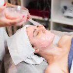 Cosmetologist applies alginate mask with spatula on face of woman. Facial skin and anti-aging treatment. Cosmetology and professional facial skin care.
